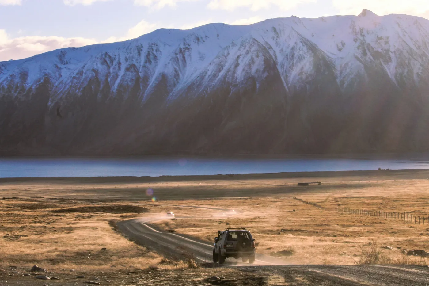 Four-wheel-drive vehicle on a dusty gravel road across golden grassland, a lake and a snow-covered mountain range behind, low evening sun