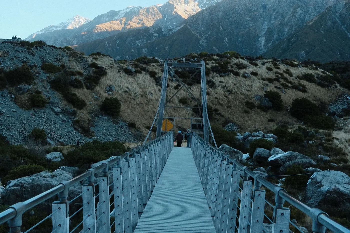 Swing bridge with metal railings and a wooden deck leading across a rocky valley towards snow-capped peaks, clear sky