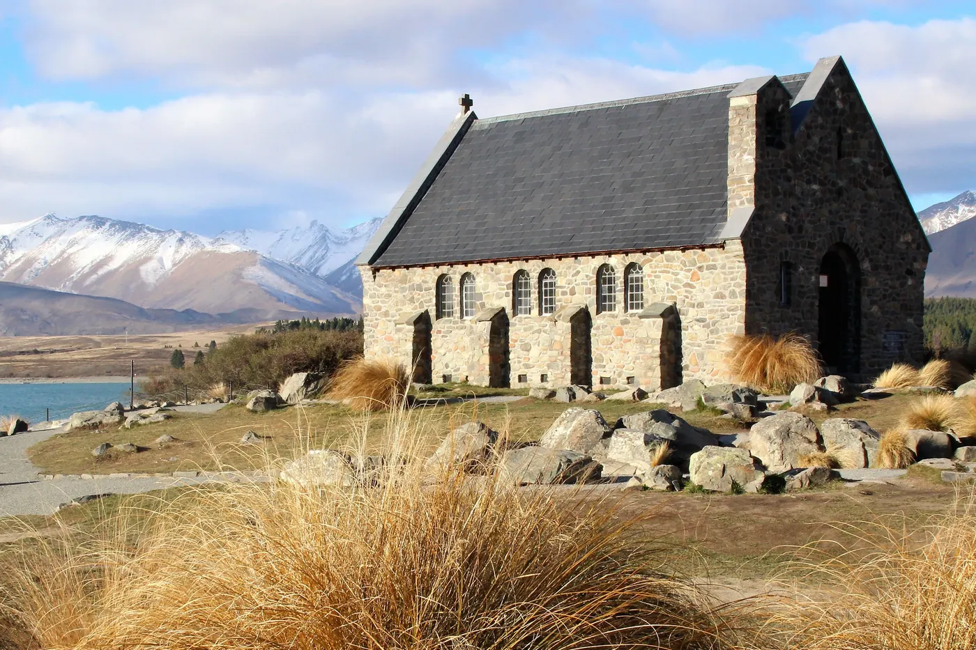 Stone Church of the Good Shepherd on the lakeshore, tussock grass and boulders in front, snow-capped mountains and blue lake behind