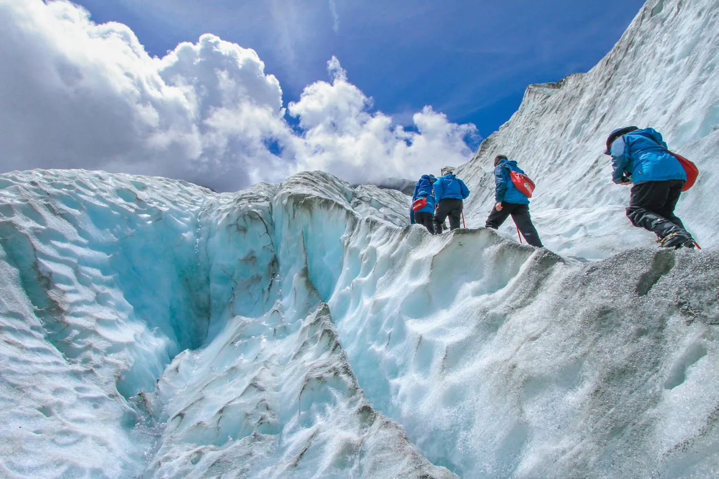 Four hikers in blue jackets climbing a ridge of blue glacier ice, seen from behind, white clouds and blue sky