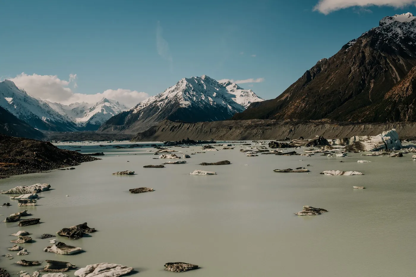 Grey glacial lake dotted with small ice floes, moraine walls and snow-capped peaks at the head of the valley