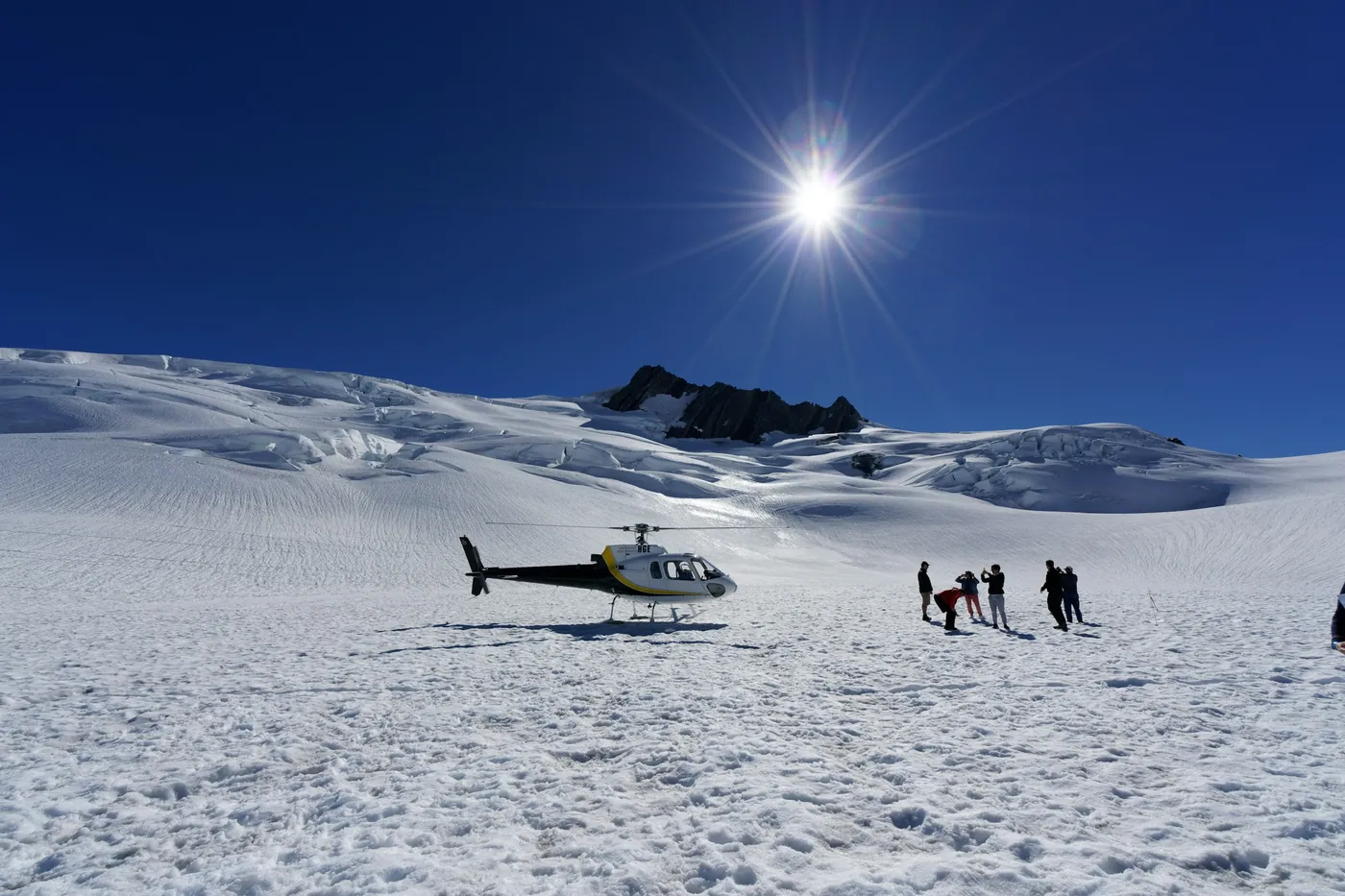 Helicopter parked on a wide snowfield, a small group of people standing beside it, bright sun in a deep blue sky, rocky peak behind