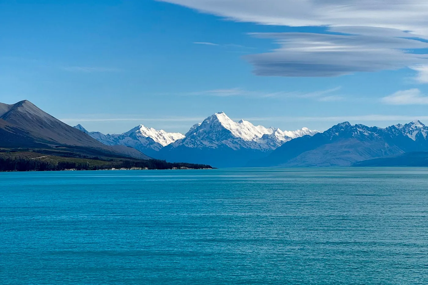 Turquoise Lake Pukaki in the foreground, Aoraki/Mount Cook and snow-capped peaks on the horizon, long lenticular clouds in a deep blue sky