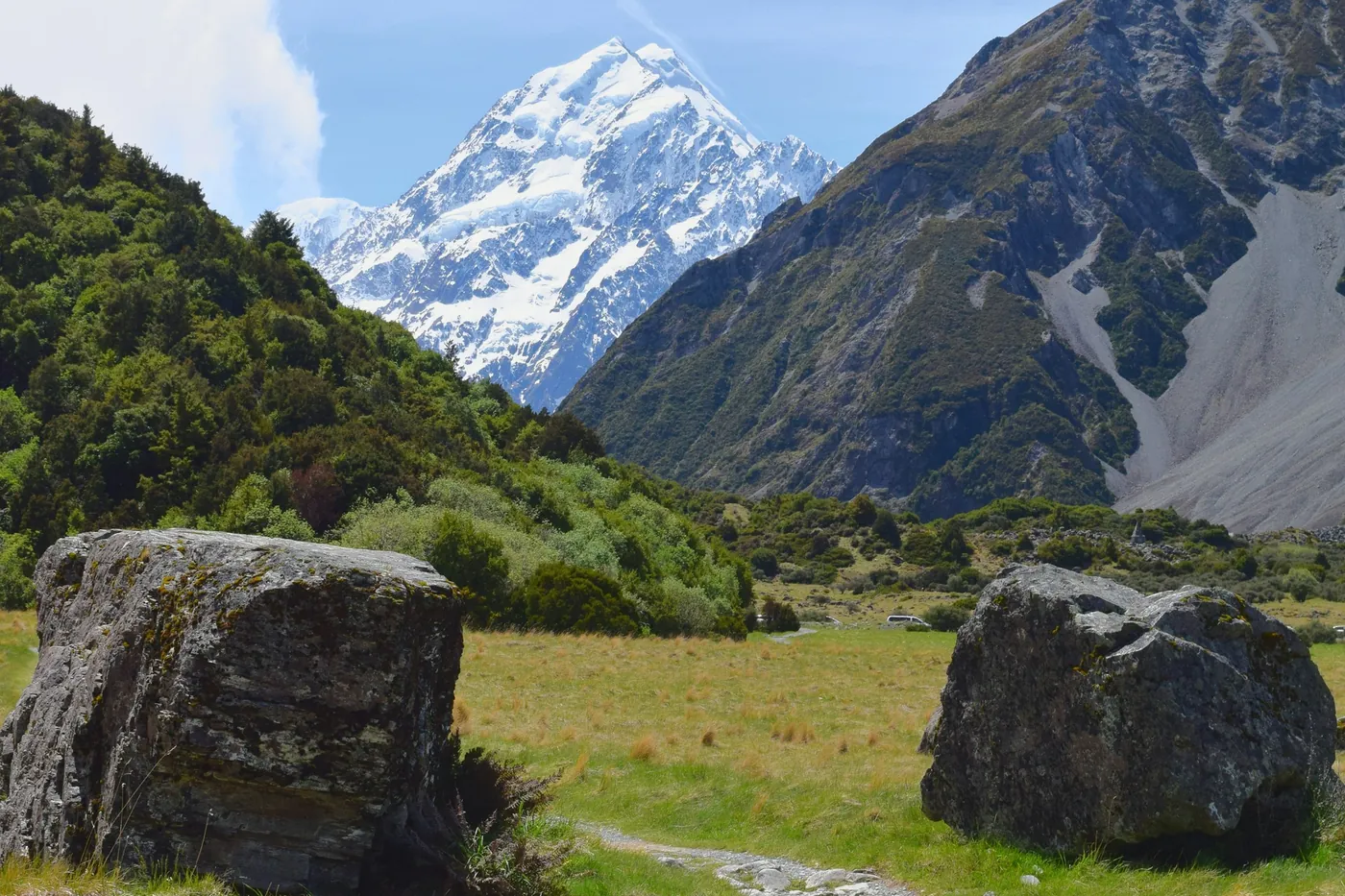 Gravel walking track between two large boulders through green grassland, bush-covered slopes and the snowy summit of Aoraki/Mount Cook behind
