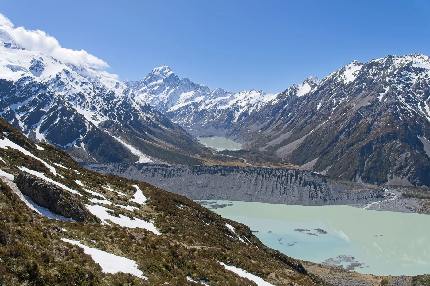 Milky glacial lake below a grey moraine wall, snowy slopes in the foreground, Aoraki/Mount Cook at the head of the valley