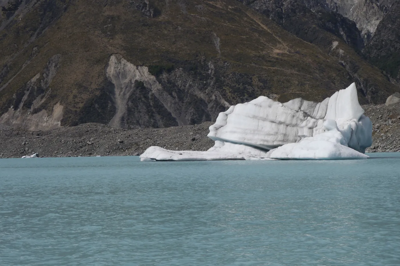 Large white iceberg floating in grey-green glacial lake water, steep moraine and mountain slope behind