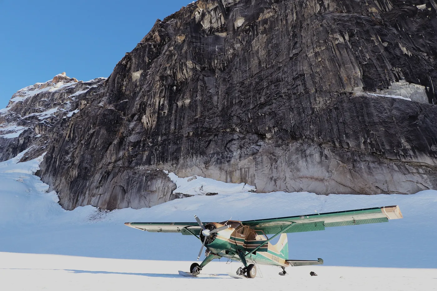 Green and cream ski-equipped bush plane parked on a snowfield below a huge dark granite rock face, clear blue sky
