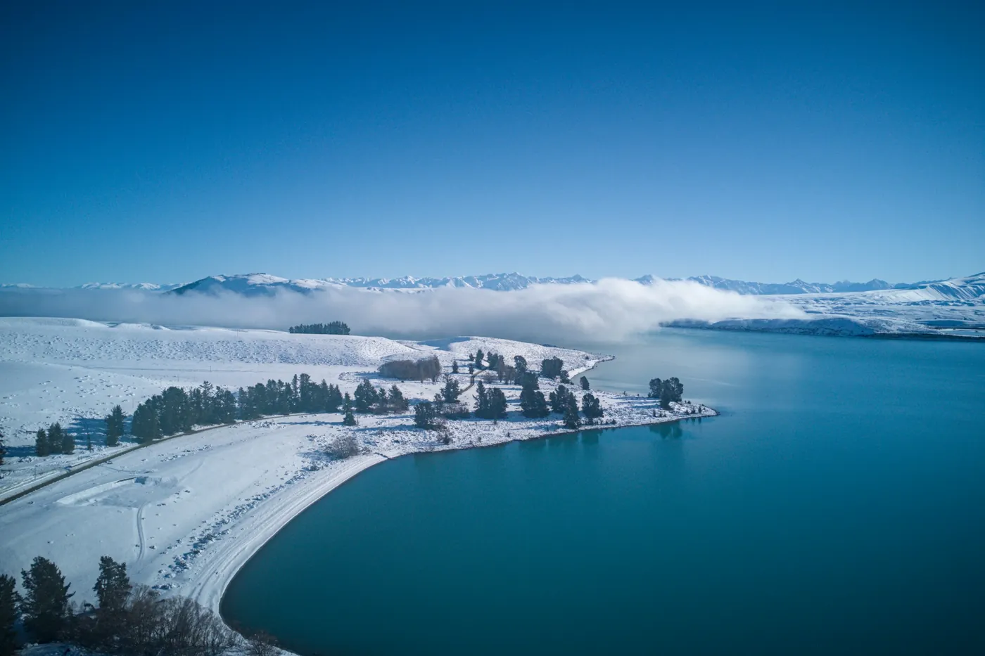 Aerial view of a snowy lakeshore with a curving road, pine trees and a deep blue lake, low fog and white hills behind
