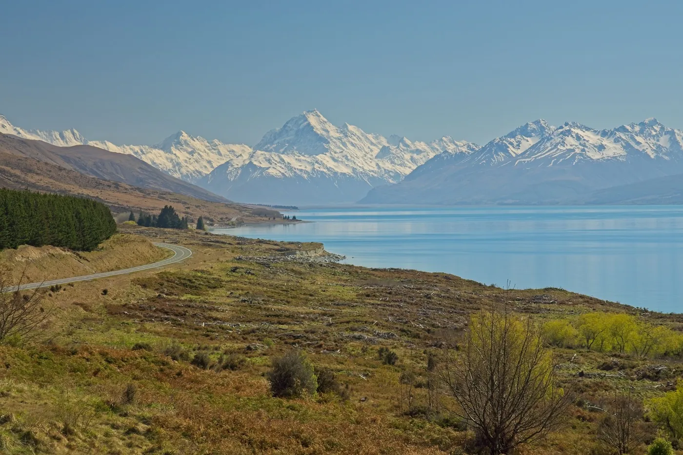Lake Pukaki stretching towards Aoraki/Mount Cook, scrubby hillside and a road curving along the shore in the foreground