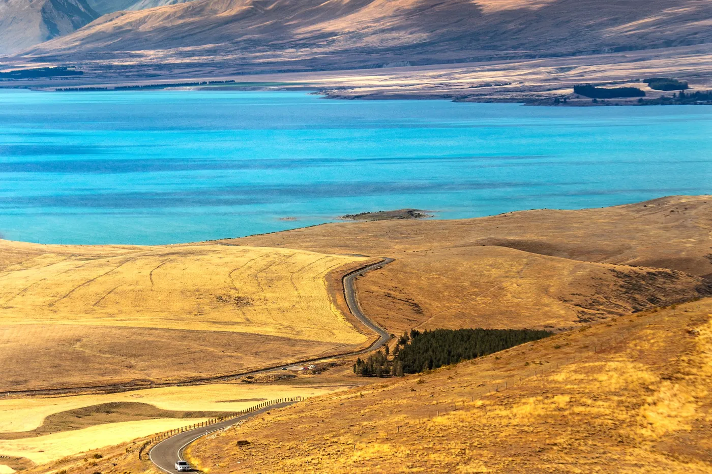 Aerial view of a winding road through golden hills above the turquoise water of Lake Tekapo, brown ranges and clouds behind