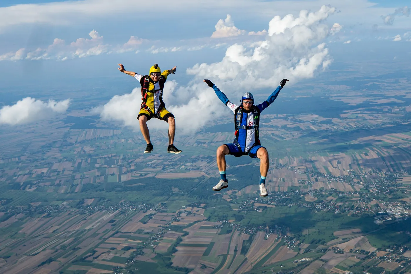 Two skydivers in free fall with arms raised above patchwork farmland and white clouds