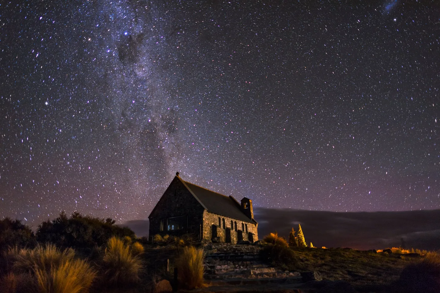 Milky Way and stars above the dark stone Church of the Good Shepherd, tussock in the foreground, faint warm lights on the horizon