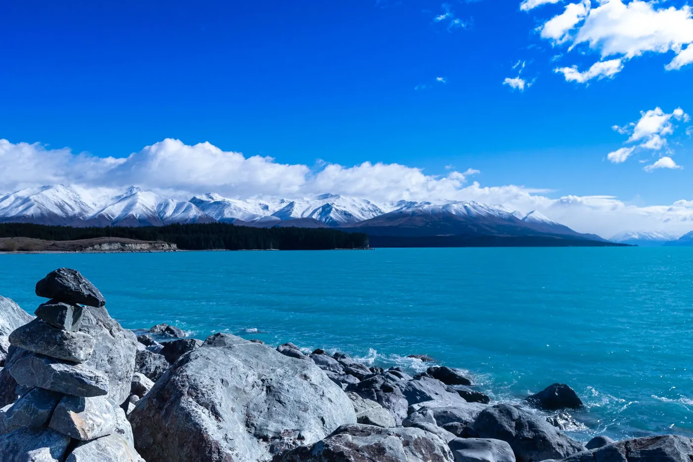 Turquoise Lake Tekapo with a stone cairn and grey boulders on the shore, snowy Southern Alps and white clouds on the horizon