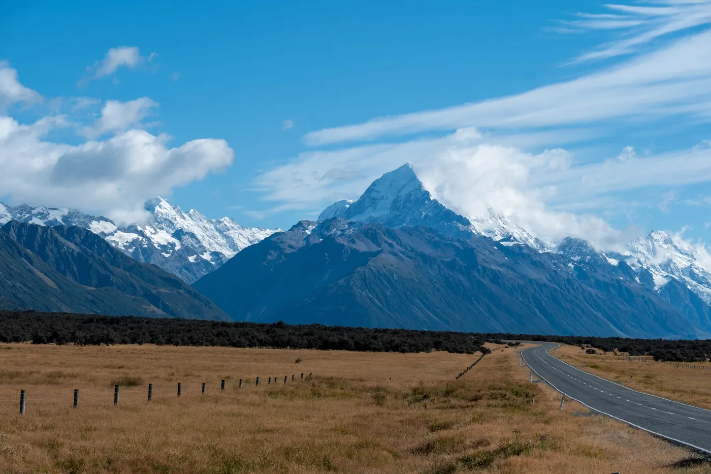 Golden tussock grassland with a fence line and a road curving towards Aoraki/Mount Cook, blue sky with clouds