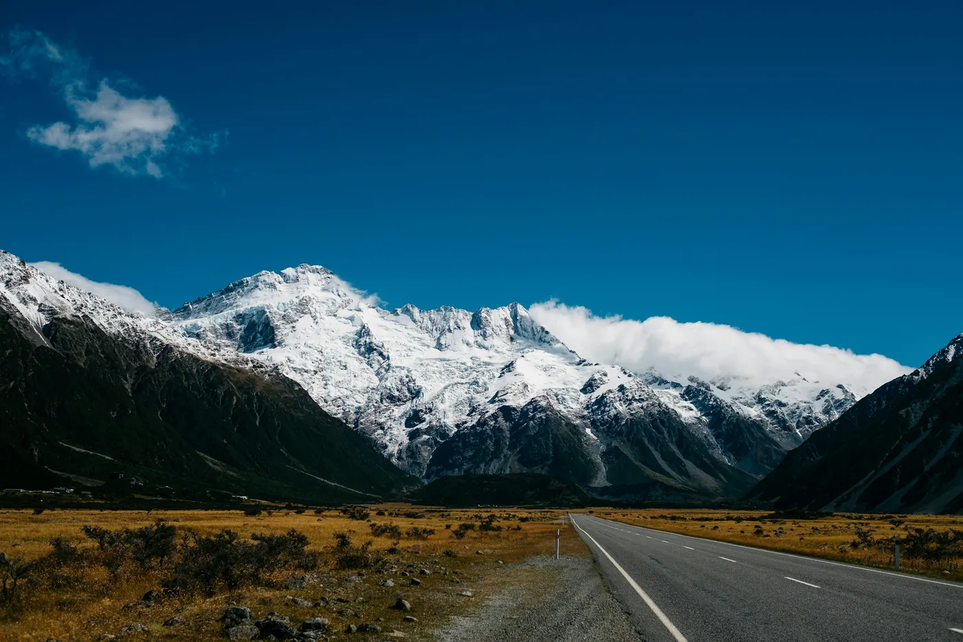 Empty road leading into a valley towards snow-capped peaks, dark slopes on both sides, tussock beside the road